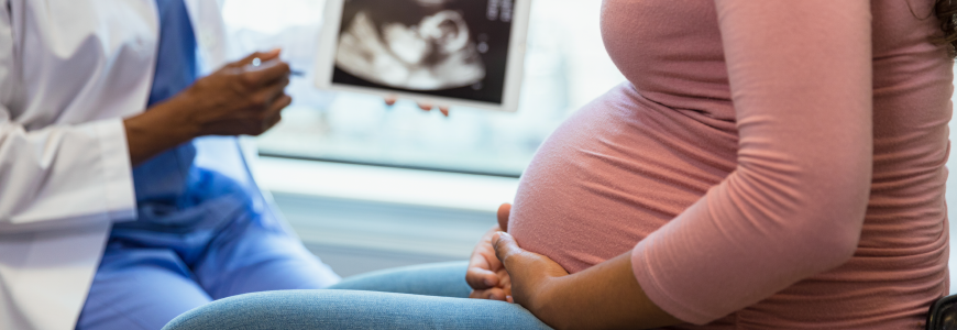 A photo with the focus on the unrecognizable pregnant woman in the foreground as the unrecognizable doctor shows her an ultrasound on a digital tablet in the background.