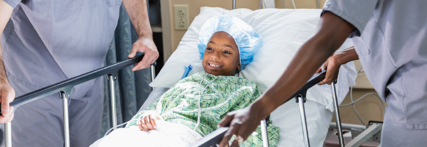 A 6 year old African-American girl in a hospital, lying on a hospital gurney wearing a surgical cap and hospital gown. A male nurse and female doctor are caring for her as she recovers from a medical procedure.