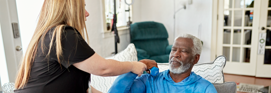 Early 70s Black man in blue tracksuit sitting in living room and doing bicep curls with dumbbells as home caregiver guides his arm.