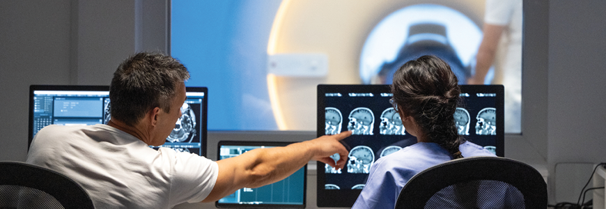 Rear view of male and female MRI technologist sitting at the console in the operating room and operating the MRI scanner
