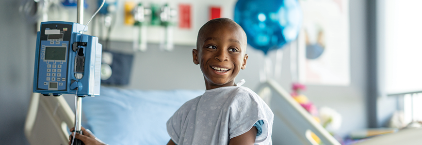 A sweet little boy of African decent, who is battling Cancer, sits on the edge of his hospital bed as he patiently waits for his Chemotherapy treatment to run. He is dressed comfortably in a gown and has a bald head from the treatments.