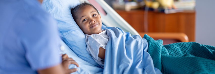 A female nurse, of African decent, makes her rounds as she checks in on a pediatric patient after surgery. The young boy is sitting up in his hospital bed and tucked warmly under the blankets as the two talk.\