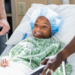 A 6 year old African-American girl in a hospital, lying on a hospital gurney wearing a surgical cap and hospital gown. A male nurse and female doctor are caring for her as she recovers from a medical procedure.