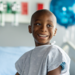 A sweet little boy of African decent, who is battling Cancer, sits on the edge of his hospital bed as he patiently waits for his Chemotherapy treatment to run. He is dressed comfortably in a gown and has a bald head from the treatments.